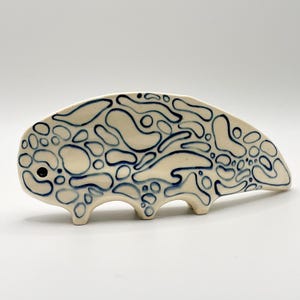 May include: A ceramic animal figurine with a cream-colored base and a hand-painted pattern of irregular dark blue organic shapes. The creature has a rounded body, a single dark blue eye, and three small legs supporting its curved, elongated form.