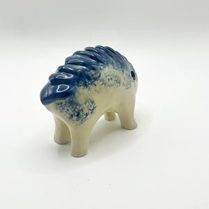 May include: A four-legged ceramic animal figurine with a smooth, cream-colored body and a textured, deep blue glaze along the back and spine. The piece features a small circular opening on the side and a ridged, segmented dorsal design.