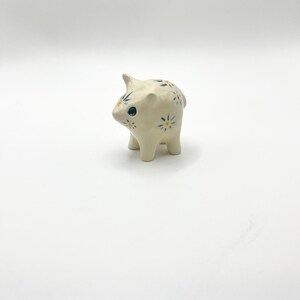 May include: A small, cream-colored ceramic pig figurine decorated with hand-painted blue and yellow daisy-like floral patterns. The pig has a smooth, glossy finish, pointed ears, and a single dark blue eye visible on the side of its head.