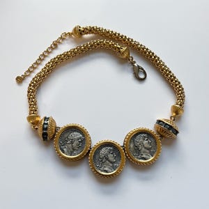 Puede incluir: Collar dorado con tres colgantes de monedas con retratos detallados. El collar tiene una cadena texturizada y detalles decorativos con piedras negras. El cierre es de tipo mosquetón.