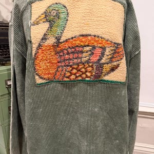 May include: A sage green long-sleeve sweatshirt with a ribbed texture and a faded wash. A large, rectangular patch featuring a textured, multi-colored goose graphic in shades of orange, yellow, green, and blue is centered on the front chest.