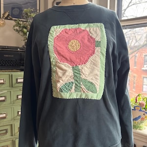 May include: A dark green sweatshirt with a large, pink and green patchwork flower design on the front. The flower is surrounded by a white border and has a yellow center.