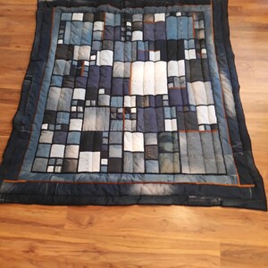 May include: A patchwork quilt made from denim fabric in shades of blue, white, and black. The quilt is made up of squares and rectangles of different sizes, creating a geometric pattern. The quilt is edged with a dark blue fabric.