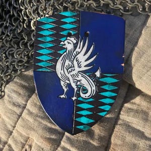 May include: A blue and teal leather shield with a white rooster design. The shield is decorated with a diamond pattern.