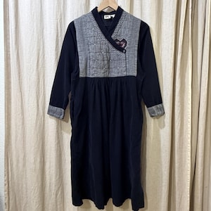 May include: A long-sleeved black dress with a grey quilted bodice and a V-neckline. The dress has a gathered waist and a small decorative element on the bodice. The sleeves have grey cuffs. The dress is hanging on a wooden hanger.