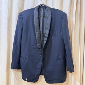 Puede incluir: Una americana azul marino con solapa de satén negro cuelga de una percha metálica. La americana tiene dos bolsillos delanteros y un cierre de un solo botón. Hay algunos agujeros pequeños cerca de la parte inferior de la americana.