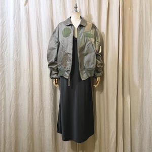 May include: A grey bomber jacket with patches and a black dress on a mannequin. The jacket has a collar, a zip, and green patches on the chest and arm. The dress is a dark grey, midi-length, and sleeveless.