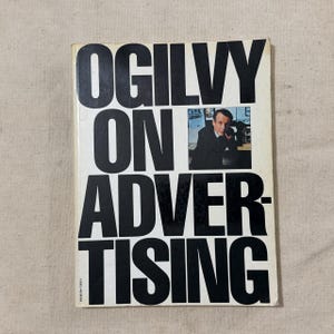 Puede incluir: Un libro titulado "OGILVY ON ADVERTISING" con texto negro grande sobre fondo blanco. Una pequeña imagen de un hombre con traje está en la esquina superior derecha. El libro está ligeramente desgastado.