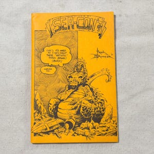 May include: A vintage comic book cover with the title "SEA-CON?!" in bold orange letters. The cover features a detailed black and white illustration of a dragon-like creature with speech bubbles. The text includes "HIC IT'S HARD TO HICE KEEP THESE 'ARTISTIC' TYPES DOWN! BURP!"