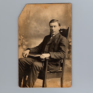 Puede incluir: Una fotografía antigua en tono sepia de una persona sentada en una silla de madera. El individuo viste una chaqueta de traje oscura y texturizada con camisa de cuello y corbata. El retrato tiene un fondo desenfocado y muestra desgaste.