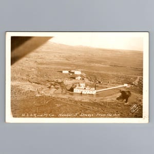 Op de afbeelding: Een sepia-kleurige vintage ansichtkaart met een luchtfoto van de U.S.S.R. and M. Co. Number 5 baggermachine. De industriële structuur staat in een landschap met een lange pijp die in een vijver uitkomt, met de tekst U.S.S.R. and M. Co. Number 5 Dredge from the Air.