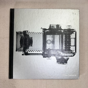 May include: A square hardcover book with a metallic silver cover featuring a black and white X-ray style illustration of a bellows camera. The title The Camera is printed in small white text in the bottom right corner of the front cover.