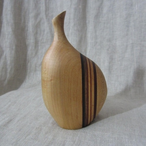 Puede incluir: Un jarrón de madera hecho a mano con un acabado marrón claro. El jarrón tiene un cuello delgado y curvado y un cuerpo redondeado. Rayas verticales de madera marrón oscuro y rojizo acentúan el lateral. El jarrón mide unos 20 cm de alto.