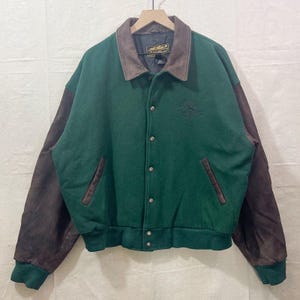 Op de afbeelding: Groene en bruine varsity jas met een bruine leren kraag en mouwen. De jas heeft een knoopsluiting, twee voorzakken en een groene geribde zoom en manchetten. Een logo is op de linkerborst geborduurd.