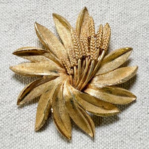 May include: A vintage gold-tone brooch in a floral design. The brooch features layered, textured petals surrounding a cluster of wheat stalks. The brooch is a bright gold color and has a pin closure on the back.