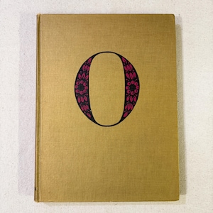May include: A vintage book with a gold cover. The cover features a large, black letter "O" filled with a pink floral design. The book's cover has a textured appearance, suggesting it is an older edition.