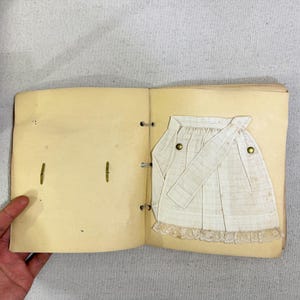 May include: An open vintage scrapbook with a cream-colored page featuring a small, cream-colored apron with a lace hem and two brass-colored buttons. The apron has a matching sash.