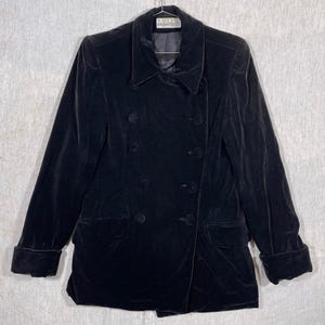 May include: A black velvet double-breasted blazer with six buttons and two front pockets. The blazer has a notched collar and long sleeves with folded cuffs. The garment is hanging on a hanger.