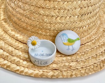 May include: A small white ceramic dish with a blue bird design and the text "20.06.2021 Mia" on the bottom. A white daisy is inside the dish. The dish is sitting on a woven straw hat.