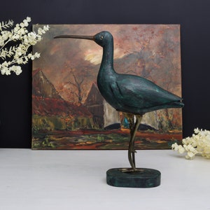 Könnte beinhalten: Eine tealblaue Holzvogelskulptur mit Messingbeinen und einem langen Schnabel. Der Vogel steht auf einem tealblauen Holzsockel. Die Skulptur steht vor einem Gemälde einer Landschaft.