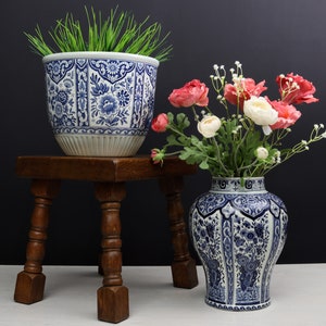 Może przedstawiać: Dwa niebiesko-białe ceramiczne wazony z kwiatowymi wzorami. Wazon po lewej stronie stoi na drewnianym stołku i zawiera zieloną trawę. Wazon po prawej stronie zawiera bukiet różowych i białych kwiatów.