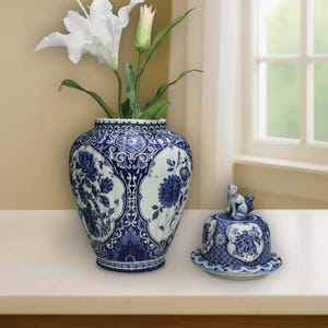 Peut inclure: Un vase en porcelaine bleu et blanc à motifs floraux contient des lys blancs. À côté, un plat couvert assorti avec une figurine de petit animal sur le dessus. Les objets sont sur une surface blanche.
