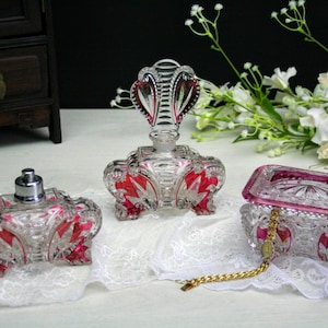 Può includere: Un set di tre flaconi di profumo vintage in vetro con accenti rosa. I flaconi sono decorati con intricati disegni in vetro tagliato e hanno un aspetto cristallino. I flaconi sono disposti su un tovagliolo di pizzo bianco.