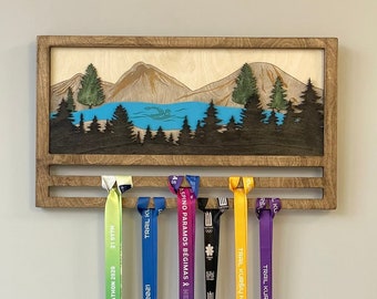 May include: Wooden medal display rack with a mountain scene and a lake. The rack has six slots for hanging medals. There are six medals hanging on the rack, with ribbons in various colors, including green, blue, purple, yellow, and pink. The medals have text on them, including "RIMI RIGA MARATHON 2020", "TRAIL KURŠIŲ NERIJA 2021", "ASPINO PARAMOS BĖGIMAS & HES MYLIME JASI", "TRAIL KURŠIŲ NERIJA 2022", and "PINK RUN".
