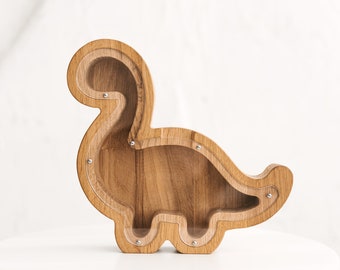 May include: A dinosaur-shaped wooden piggy bank featuring a clear acrylic front panel secured with small metal screws. The natural wood grain is visible throughout the frame, which is shaped like a long-necked sauropod dinosaur with a hollow center.