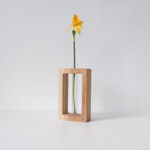 May include: A single yellow daffodil flower in a clear glass test tube vase. The vase is held in a wooden frame.