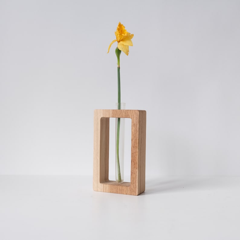 May include: A single yellow daffodil flower in a clear glass test tube vase. The vase is held in a wooden frame.