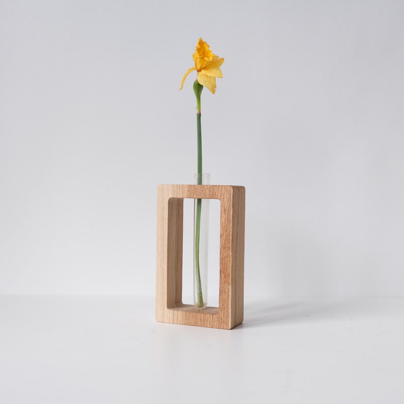 May include: A wooden frame with a glass test tube inside, holding a single yellow daffodil flower.
