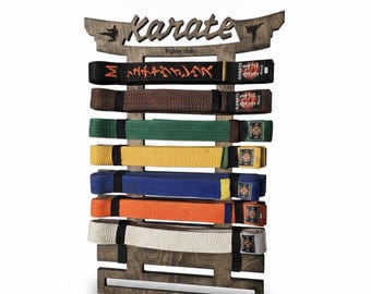 Op de afbeelding: Een donkerbruin houten wandrek voor vechtsportbanden. Bovenaan staat het woord Karate in een gestileerd lettertype met kleine silhouetten van vechters. Het rek bevat zeven gevouwen banden in zwart, bruin, groen, geel, blauw, oranje en wit.
