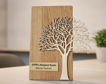 Puede incluir: Una placa rectangular de madera marrón claro con una silueta de árbol de madera clara cortada con láser en la parte frontal. Una pequeña placa metálica dorada está fijada en la parte inferior con el texto GPFB's Deepest Roots Marcia Paulson.