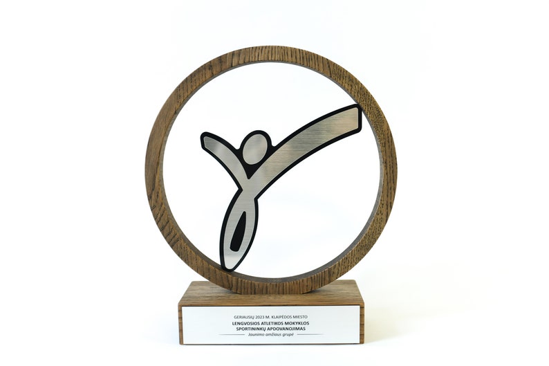 May include: A wooden award with a silver stylized figure of a person with arms raised in the air. The figure is inside a wooden circle. The base of the award has a plaque with the text "GERIAUSIU 2023 M. KLAIPĖDOS MIESTO LENGVOSIOS ATLETIKOS MOKYKLOS SPORTININKŲ APŽVALGINIMAS  -  &bdquo;Sportinis andrius grupė"