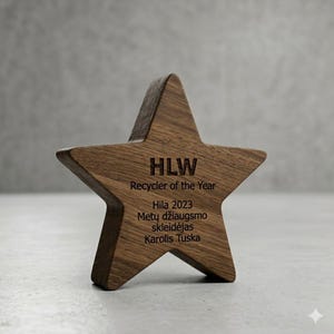 Op de afbeelding: Een stervormige houten onderscheiding met een donkerbruine houtnerf. De voorzijde is gegraveerd met de tekst: HLW, Recycler of the Year, Hila 2023, Metų džiaugsmo skleidėjas, Karolis Tuska. Het object heeft een dikke, platte voet om rechtop te staan.
