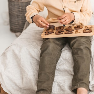 Puede incluir: Un tablero de rompecabezas num&eacute;rico de madera con n&uacute;meros recortados en color marr&oacute;n oscuro del cero al nueve, encajados en una base de madera clara. Las piezas tienen forma de d&iacute;gitos individuales para colocar en sus ranuras.