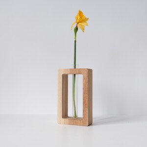 May include: A wooden vase with a clear glass tube for holding a single flower. A yellow daffodil is in the vase.