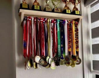 May include: A light-colored wooden wall-mounted shelf featuring a flat top surface holding six gold-colored trophies with black bases. Below the shelf, a horizontal bar displays numerous hanging medals attached to colorful fabric ribbons of various patterns.