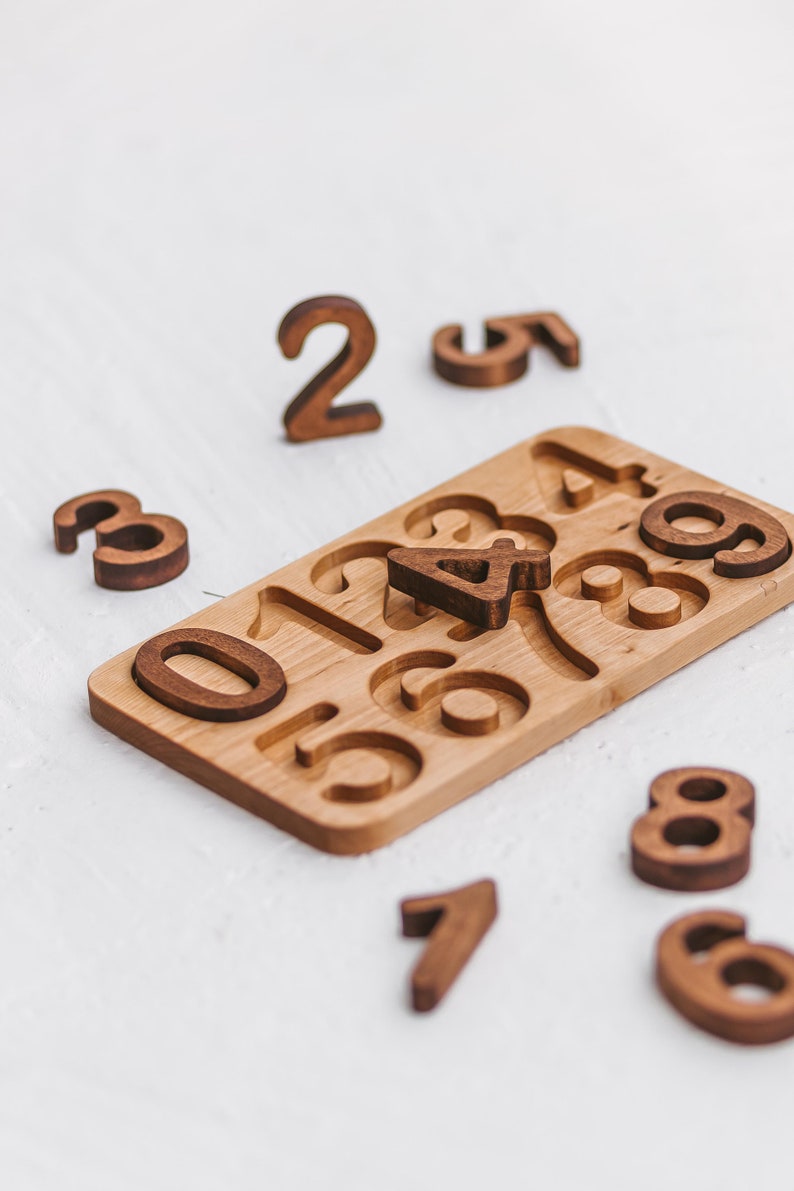 Puede incluir: Un tablero de rompecabezas rectangular de madera con recortes para los d&iacute;gitos del cero al nueve, con varias piezas num&eacute;ricas de madera marr&oacute;n oscuro esparcidas alrededor y el n&uacute;mero cuatro colocado en su ranura correspondiente.