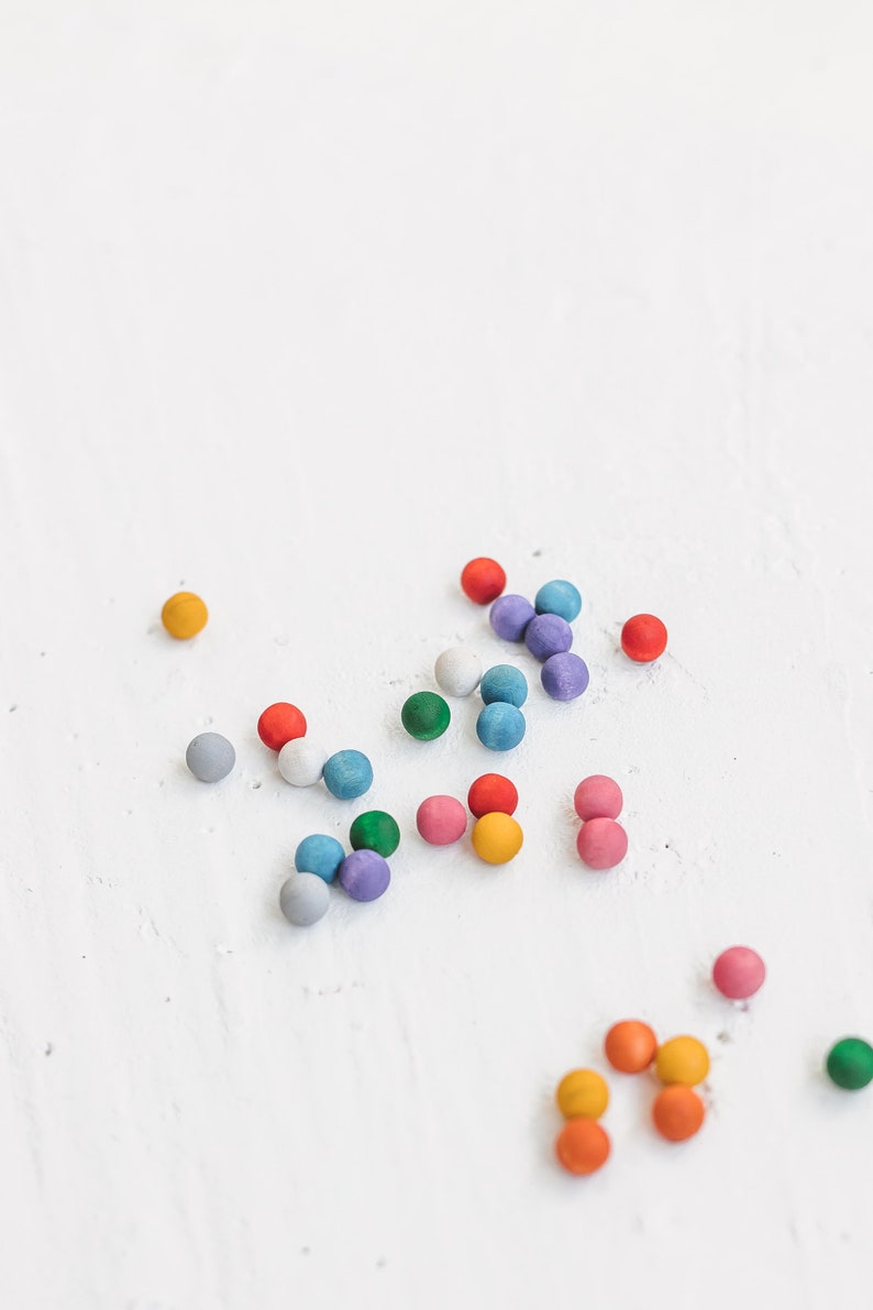 May include: A collection of colorful wooden balls in various shades of blue, green, red, orange, yellow, and purple scattered on a white surface.