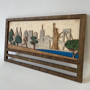 Puede incluir: Un expositor de medallas de pared de madera con un diseño de corte por láser del horizonte de la ciudad de Nueva York, que incluye la Estatua de la Libertad, el puente de Brooklyn y un corredor. El expositor tiene dos barras horizontales para colgar medallas.