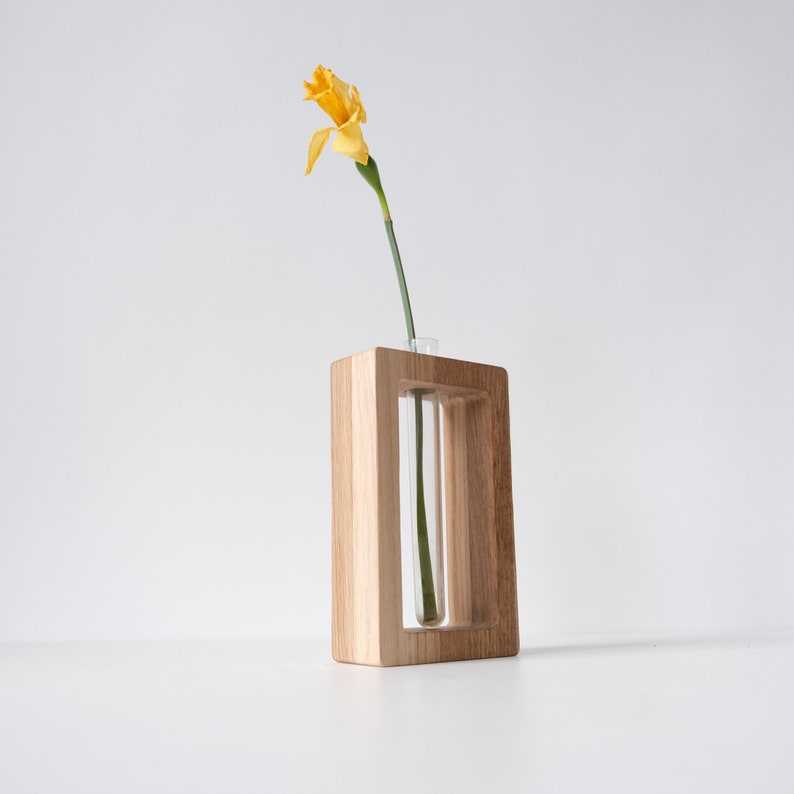 May include: A wooden vase with a clear glass tube for holding a single flower. The vase is light brown and has a rectangular shape. A yellow daffodil is in the vase.