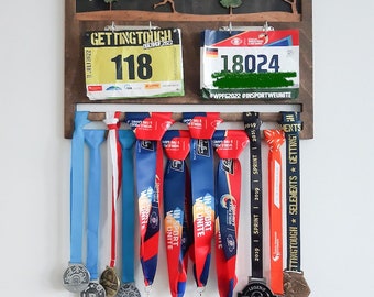 May include: A wooden wall-mounted display rack featuring a forest silhouette at the top, two race bibs labeled 118 and 18024, and multiple colorful fabric ribbons with attached metal medals hanging from the bottom bar in various designs.