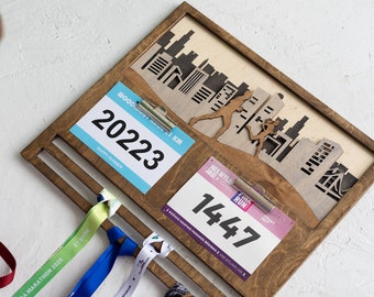 May include: A rectangular wooden display board with a dark brown finish, featuring two metal clips holding race bibs numbered 20223 and 1447. The top section displays a laser-cut wooden cityscape silhouette with running figures. Four colorful race medals hang from slots.