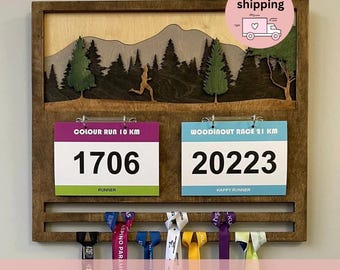 May include: A wooden race medal display rack featuring a layered mountain and forest scene with a runner silhouette. It includes two clips holding race bibs labeled Colour Run 10 KM 1706 and Woodinout Race 21 KM 20223, with hanging ribbons below.