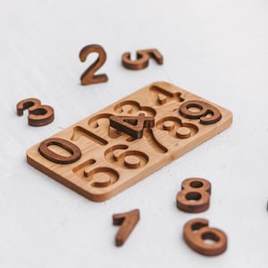 Puede incluir: Un tablero rectangular de madera con recortes para los n&uacute;meros del cero al nueve, con varias piezas num&eacute;ricas de madera marr&oacute;n oscuro esparcidas alrededor, incluido un n&uacute;mero cuatro colocado en el centro del tablero.