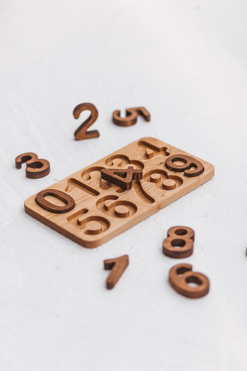 Puede incluir: Un tablero rectangular de madera con recortes para los n&uacute;meros del cero al nueve, con varias piezas num&eacute;ricas de madera marr&oacute;n oscuro esparcidas alrededor, incluido un n&uacute;mero cuatro colocado en el centro del tablero.