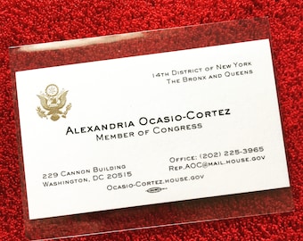May include: A white business card with gold lettering. The card reads "14th District of New York The Bronx and Queens Alexandria Ocasio-Cortez Member of Congress 229 Cannon Building Washington, DC 20515 Office: (202) 225-3965 Rep.AOC@mail.house.gov Ocasio-Cortez.house.gov". The card also features a gold seal of the United States.