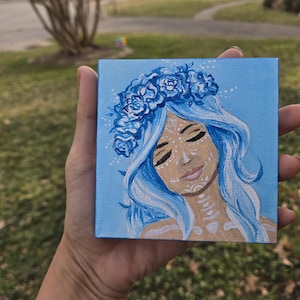 May include: A small square canvas painting depicting a woman with blue hair and a floral crown. The artwork has a light blue background and the woman's face has white decorative markings. The painting is held in a hand.